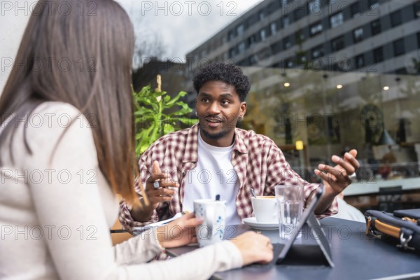 African american man and caucasian woman in lively outdoor cafe meeting, sharing ideas over coffee while reviewing a tablet, collaborating and networking in an urban setting