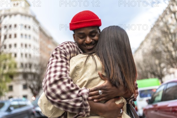 Young man in a red beanie smiles as he embraces his partner on a city street, sharing a warm, candid moment of affection, connection and genuine joy between two people