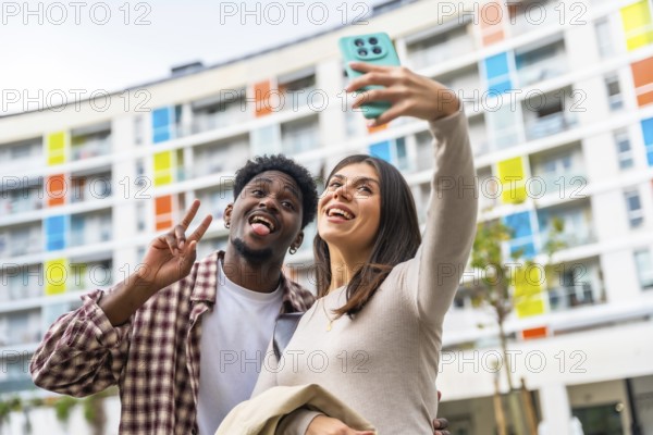 Multiethnic couple enjoying leisure time together, standing outdoors in front of a colorful modern building, playfully posing for a selfie using a mobile phone