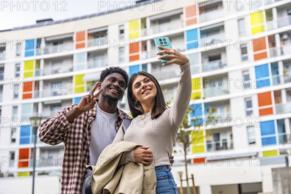 Happy diverse couple smiling and posing for a smartphone selfie, showing a peace sign while standing outdoors in front of a modern residential building with vibrant balconies