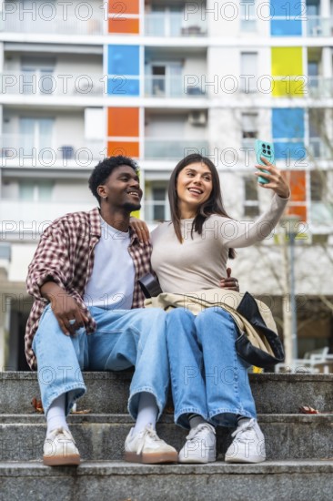 Happy diverse young couple sitting on concrete steps, smiling and taking a cheerful selfie together with a smartphone in a modern city setting with colorful buildings in the background