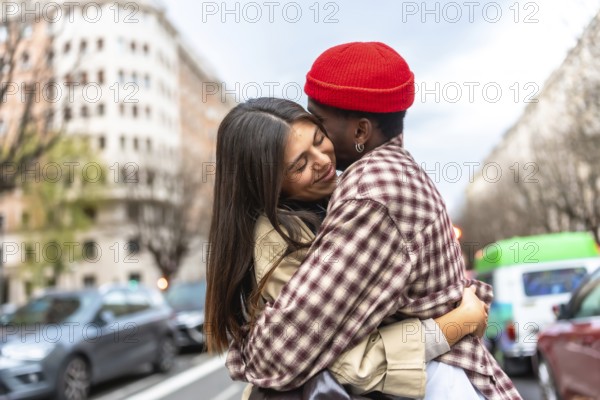 Young diverse couple embracing on an urban street, expressing love and affection, with a multi ethnic man kissing the smiling woman's cheek while surrounded by city buildings and cars