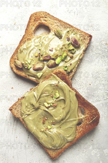 Two slices of toasted bread, topped with pistachio paste and sprinkled with pistachios, spread on a light surface
