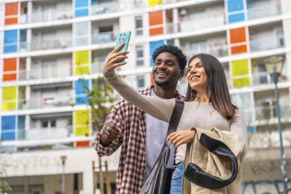 Happy young multiracial couple smiling, taking a selfie with a mobile phone in a vibrant urban environment with a colorful modern building background, enjoying city life and social media