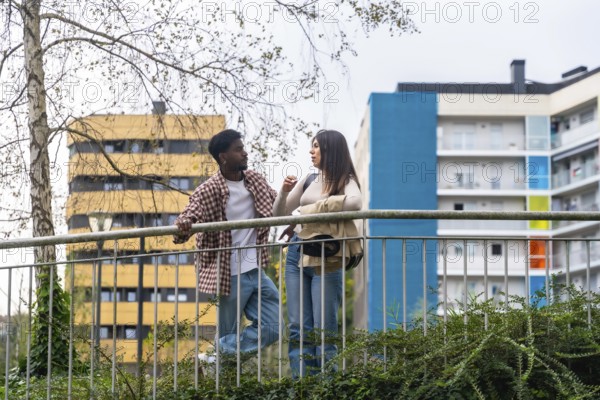 Two young adults, african american man and caucasian woman, chatting and leaning on a metal railing on a city street with buildings and green foliage behind them, urban friendship