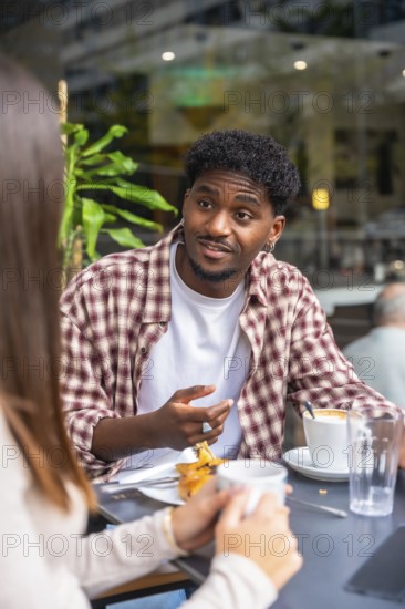 Young black man engaging in a lively conversation with a woman at an outdoor cafe table, enjoying coffee and pastries during a relaxed social gathering