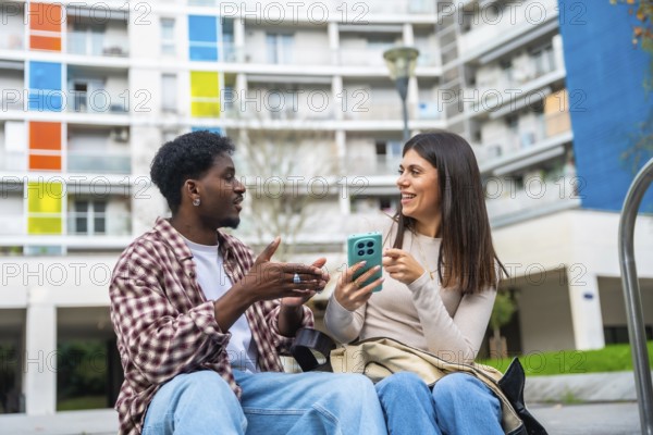 Diverse friendship bond as a young african american man and a young caucasian woman sit outdoors, sharing a laugh and looking at a smartphone in an urban setting