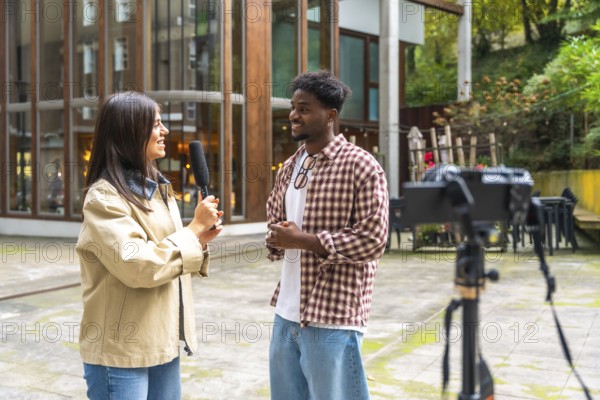 Female reporter holding a microphone, interviewing a smiling young african american man outdoors while a video camera records the interaction on a tripod, capturing live content
