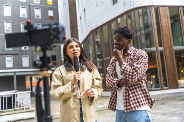 Female journalist holding a microphone and gesturing while interviewing a young man on a city street, documenting current events for a live news broadcast