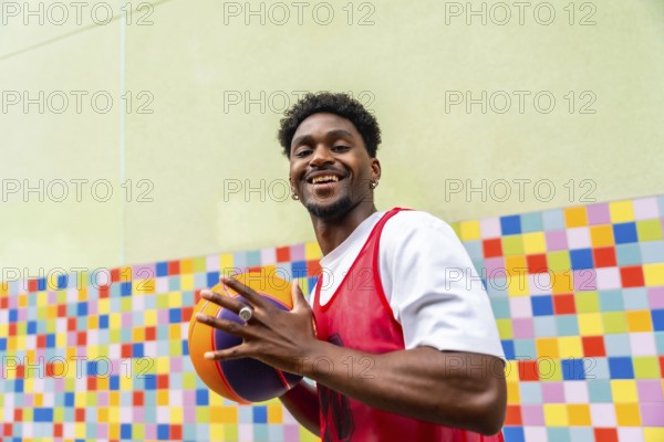 Young man smiling and holding a vibrant basketball in sportswear, standing against a colorful tiled wall outdoors urban streetball vibe, active lifestyle and joy