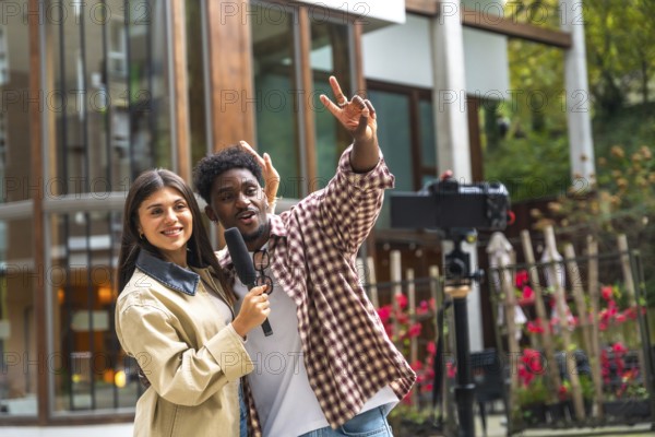 Diverse woman holding a microphone and smiling while man gestures cheerfully as they film a vlogging video with an external camera on a tripod in an urban outdoor setting
