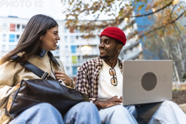 Diverse young students sitting outdoors in an urban environment, engaging in conversation and smiling while one uses a laptop, representing connection and collaboration