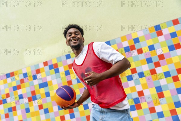 Young black man happily holding a basketball, dressed in a red sports vest and jeans, posing against a vibrant, mosaic tiled background, symbolizing urban youth and active lifestyle