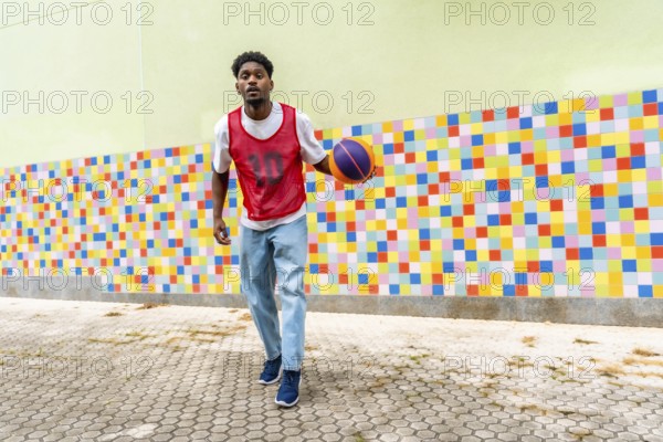 Young black man in casual sportswear playing and dribbling a basketball on a paved court against a vibrant, multi colored wall, representing youth, sport, and urban lifestyle