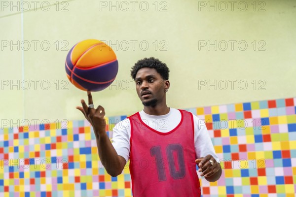 Young black man in sportswear displaying impressive skill while balancing a colorful basketball on his finger, focusing intently against a vibrant, playful background