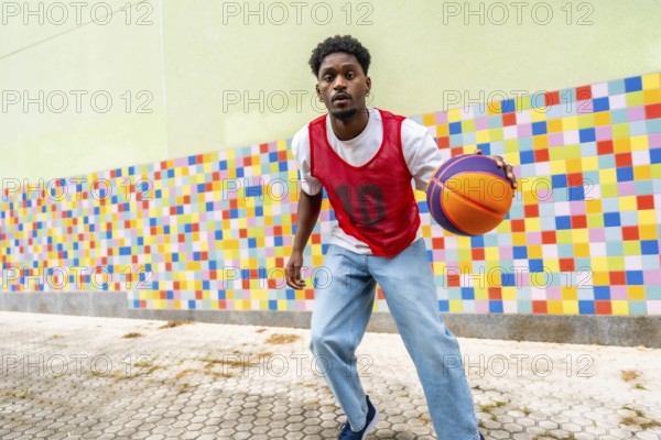 Young man actively playing street basketball, dribbling the ball with focus and energy against a vibrant, mosaic patterned wall, embodying urban sport and active lifestyle