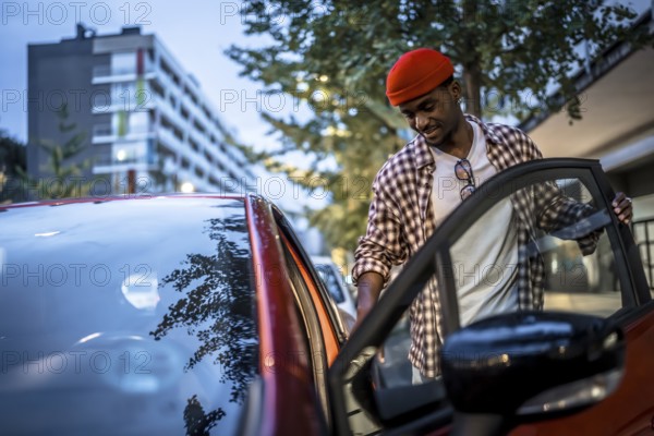 Young black man in a red beanie smiling as he opens a car door on a city street, ready to drive through an urban neighborhood with buildings and trees nearby