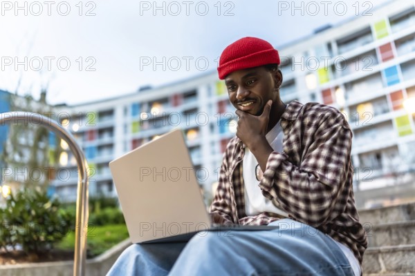 Young black man sitting on apartment steps with laptop, smiling and thoughtful as he works or studies outdoors, embodying modern urban remote work and digital lifestyle