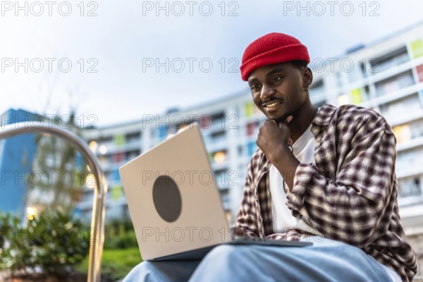 Young black man smiling while working on a laptop computer with casual clothes and a red beanie, sitting outdoors in an urban environment with modern buildings in the background