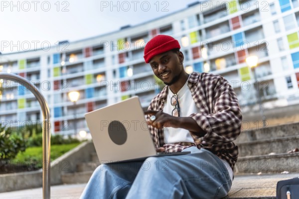 Young black man in casual clothing using a laptop while sitting on steps in an urban setting, enjoying remote work and digital nomad lifestyle with a colorful modern building in the background