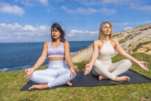Two healthy multiracial women performing yoga and meditation poses in lotus position on a yoga mat during a mindful wellness activity outdoors by the ocean in nature