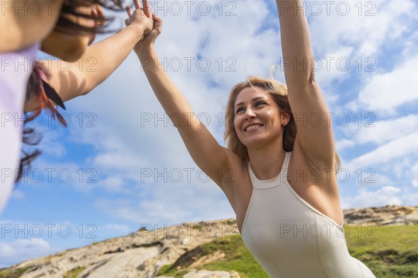 Two smiling women stretching their arms to the sky, connecting their hands in a yoga pose outdoors, symbolizing friendship, wellness, and self care in a natural landscape
