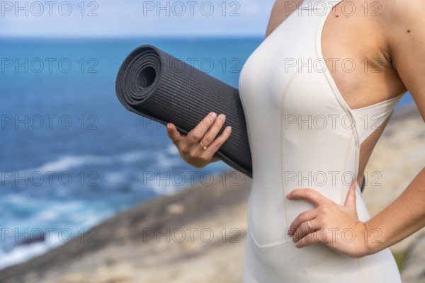Woman standing outdoors holding a rolled yoga mat, preparing for a wellness and fitness session by the ocean, connecting with nature and an active, healthy lifestyle