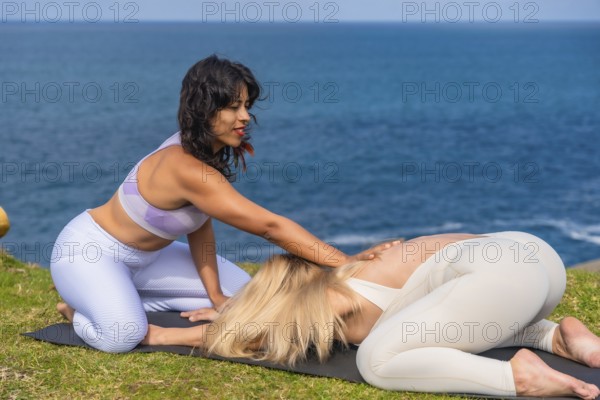 Instructor guiding woman in child's pose on grassy seaside during outdoor yoga session, promoting relaxation, mindfulness and well being with calm ocean backdrop