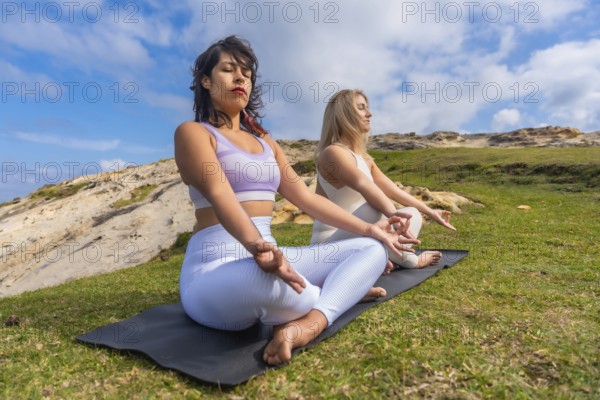 Two young women meditating on yoga mats outdoors, finding peace and mindfulness in nature, practicing spiritual wellness and healthy lifestyle under a blue sky