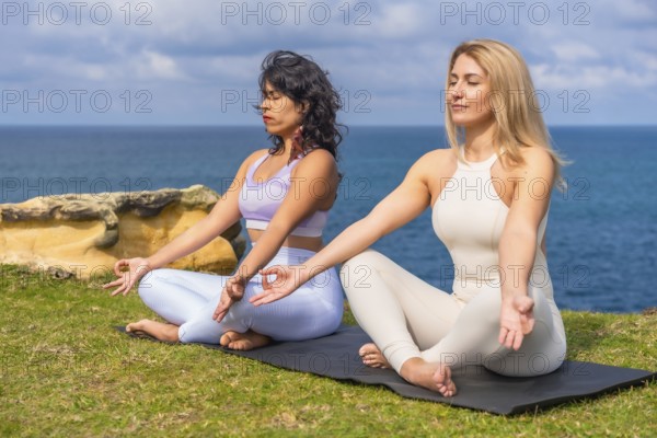 Two diverse women sitting in lotus pose on yoga mats by the ocean, finding tranquility and balance through mindful meditation for wellness and spirituality