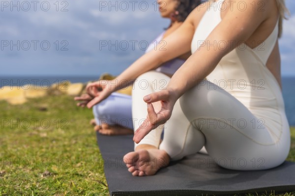 Two women practicing yoga and meditation in lotus position on a mat, finding balance and relaxation outdoors on a grassy cliff overlooking the ocean under a clear sky