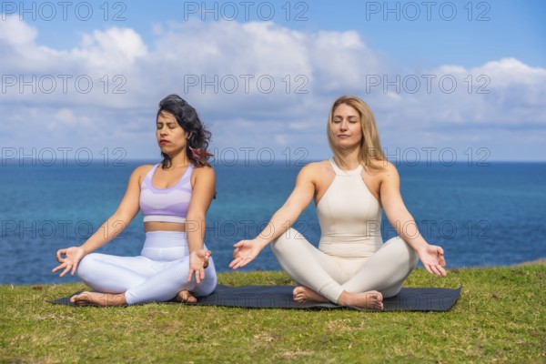 Two women sit cross legged on a mat by the ocean, practicing yoga and meditation together at sunrise, finding calm, balance and mindful connection with nature and each other