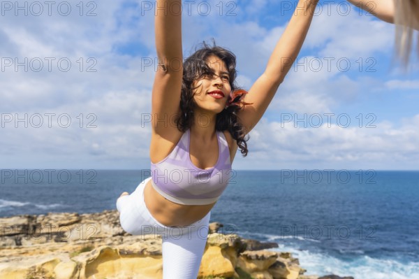 Woman balancing in a yoga warrior pose, stretching arms towards the sky on a cliff overlooking the blue ocean under a bright, cloudy sky, symbolizing wellness and freedom