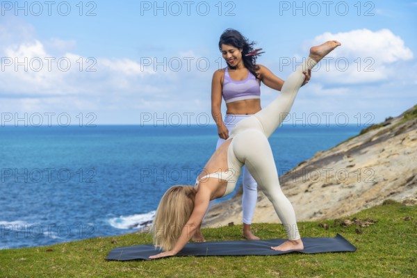 Two women stretching on a yoga mat during an outdoor yoga class by the beautiful blue ocean, focusing on flexibility and balance with guidance from an instructor
