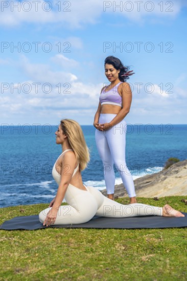 Two women stretching on a yoga mat by the ocean while practicing yoga and meditation, enjoying a serene healthy active lifestyle on a sunny day during a wellness retreat