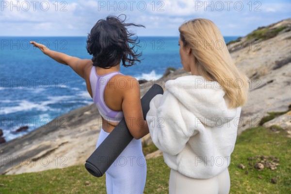 Two women standing together on a grassy cliff overlooking the ocean, one raising an arm and holding a yoga mat while looking at the sea, preparing for a mindful exercise session