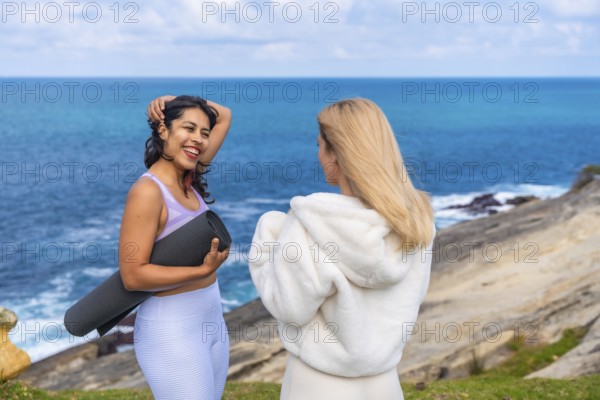 Two women friends standing on a cliff overlooking the ocean, one holding a yoga mat and laughing while talking with the other, enjoying a wellness lifestyle
