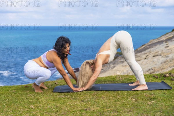 Two women practicing yoga with an instructor correcting posture during a wellness session outdoors on a grassy cliff overlooking the blue ocean, promoting health and fitness