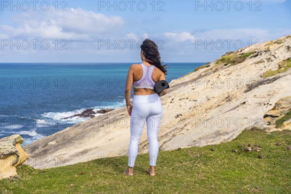 Woman standing on a grassy cliff edge, holding a yoga mat and looking out at the vast blue ocean, embracing wellness, peace, and an active lifestyle in nature