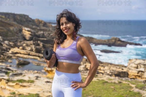 Woman standing on a rocky coastline holding a yoga mat, preparing for an outdoor fitness session with the ocean and sky creating a serene backdrop for health and wellness activities