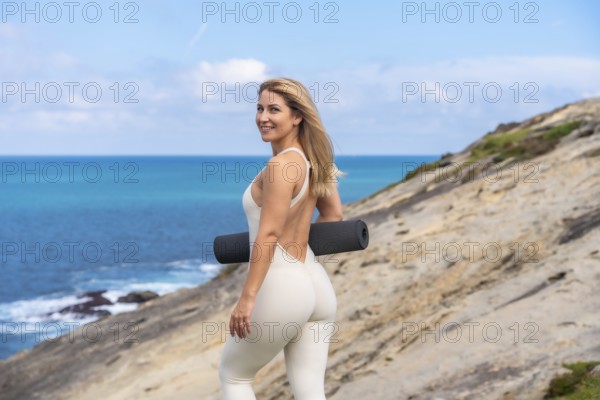 Woman holding a yoga mat, ready to practice mindfulness and engage in a healthy lifestyle with fitness activities and meditation on a coastal cliff with the vibrant blue sea in the background