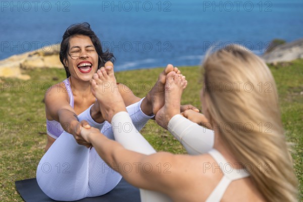 Two women performing a yoga pose together on a mat on green grass by the sea, connecting through shared laughter and exercise, promoting friendship and wellness in nature