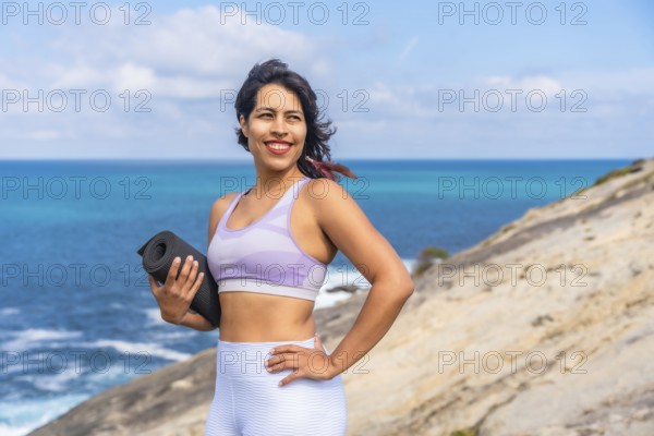 Woman smiling, holding a rolled yoga mat on a rocky cliff, looking towards the bright blue ocean, representing a healthy lifestyle, fitness, and wellness journey