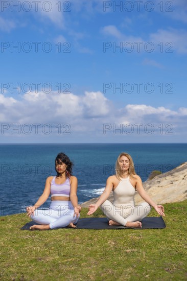 Two women sitting on yoga mats in a lotus position with eyes closed, finding tranquility and balance through yoga and meditation practice outdoors by the ocean