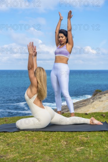 Women practicing yoga poses and meditation outdoors, an instructor guiding a student on a yoga mat with the blue ocean and sky creating a serene background