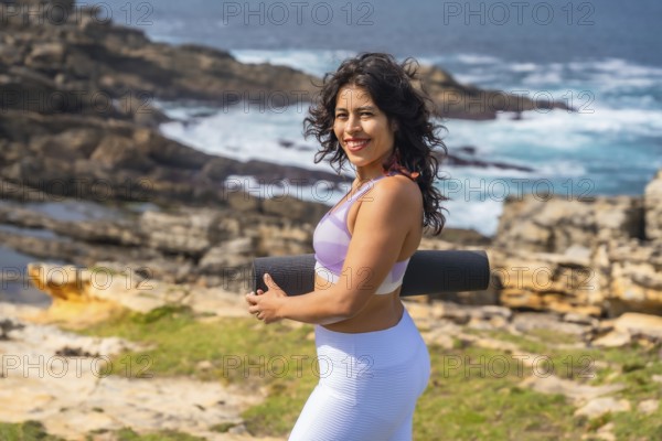 Woman smiling and carrying a rolled yoga mat or exercise mat, standing on a rocky coastline with green vegetation and the ocean in the background, embracing health and wellness outdoors