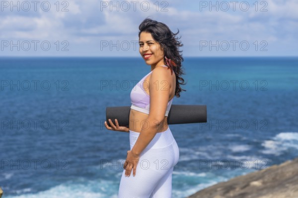 Woman smiling and holding a black yoga mat, standing on a cliff edge with the vast blue ocean and sky creating a serene background, ready for wellness and fitness