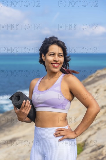 Woman holding a yoga mat and smiling, standing on a rocky cliff overlooking the blue ocean during daytime, enjoying an active and healthy lifestyle while practicing yoga and meditation in nature