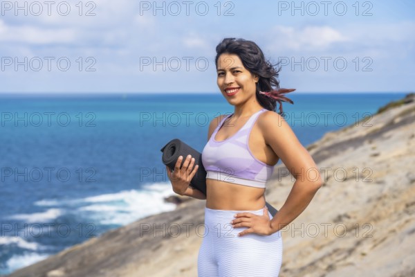 Woman smiling and holding a rolled yoga mat, standing confidently on a coastal cliff overlooking the vast blue ocean on a sunny day, representing wellness, mindfulness, and an active lifestyle