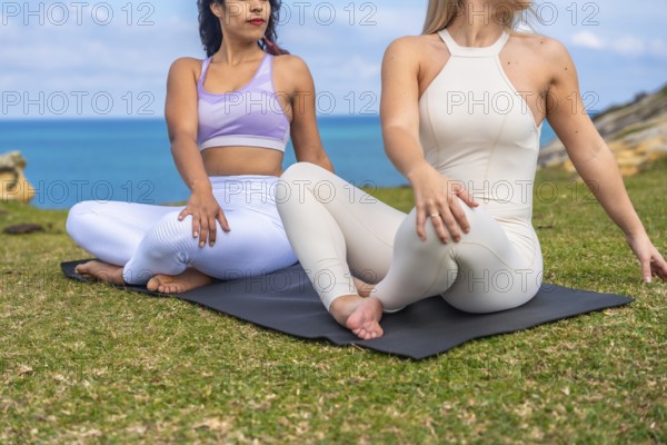 Two diverse women in seated spinal twist yoga poses on mats, meditating and connecting with nature by the ocean under a clear sky, calm and serene seaside wellness session